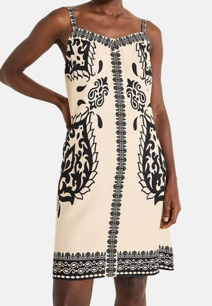 Beżowa sukienka bez rękawów z czarnymi symetrycznymi wzorami paisley i kwiatowymi, kwadratowym dekoltem i cienkimi ramiączkami, noszona przez stojącą osobę.