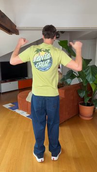 Hombre de pie en la sala señalando con los pulgares la parte trasera de una camiseta amarilla "Santa Cruz" con gráfico de palmera, vistiendo vaqueros azules y zapatos blancos.