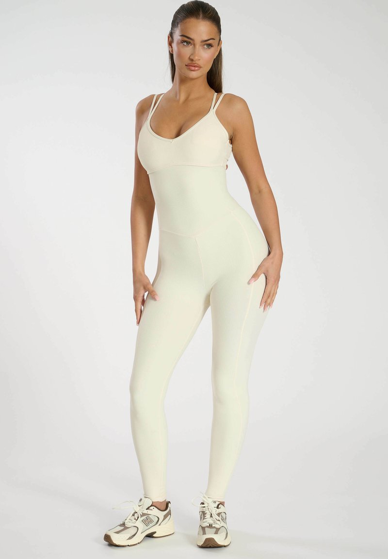 Krämfärgad ribbad jumpsuit med tunna band, figurnära silhuett och kontrasterande sömmar. Parat med neutrala träningsskor.