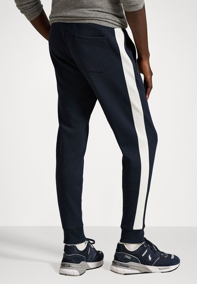 Jūras zila jogger stila bikses ar baltu sānu joslu, pieguļošu piegriezumu un aizmugures kabatu. Saskaņotas ar tumšiem sporta apaviem. Gluda kokvilnas un sintētisko šķiedru auduma maisījums.