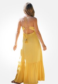Gele maxi-jurk met een gerimpelde zoom, spaghettibandjes en een open rug. Kenmerkt zich door franjes op de taille en een luchtige, lichte stof.