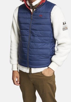 Person iført en blå quiltet vest over en hvid fleecejakke med broderede logoer og brune bukser, stående med den ene hånd i lommen.