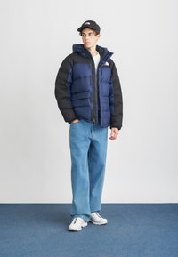 Puhasta jakna u mornarsko plavoj i crnoj boji, s kapuljačom i više džepova sa patentnim zatvaračima. Plave traperice širokih nogavica i bijele sportske tenisice upotpunjuju izgled.