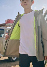 Beige leichte Jacke mit Kapuze, ausgestattet mit einem hellgelben Netzfutter und einem grünen Reißverschluss, getragen über einem schlichten weißen T-Shirt.