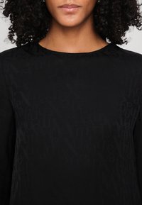 Femme aux cheveux bouclés portant un haut noir avec un motif texturé subtil de lettres éparses.