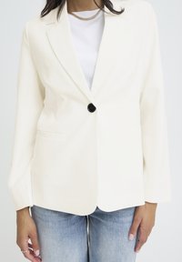 Blazer blanc avec un seul bouton noir, revers crantés et deux poches avant. Superposé sur un t-shirt blanc uni. Design classique.