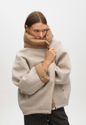 Chaqueta de mezcla de lana color beige con un cuello acolchado en tono marrón claro y detalles en cremallera dorada. Presenta mangas anchas y un ajuste relajado. Tejido texturizado.