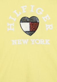 Gul bomulds T-shirt med et hjerteformet paletdesign med røde, hvide og blå sektioner samt "HILFIGER NEW YORK" tekst ovenfor og nedenfor.