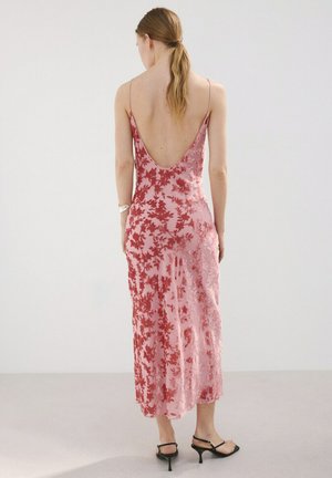 Femme aux cheveux blonds attachés en queue de cheval basse, portant une robe nuisette à fleurs roses et rouges avec un dos décolleté et des sandales à talons noires.