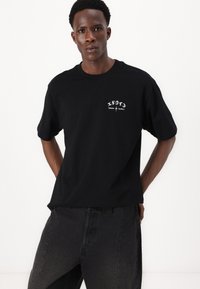 T-shirt in cotone nero con maniche corte, caratterizzato da testo grafico bianco e logo sul petto. Abbinato a pantaloni in denim grigio scuro.