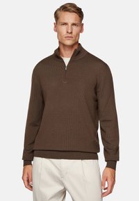Brown zip-up sweater with ribbed texture and a high collar. Features long sleeves and a relaxed fit, paired with light-colored trousers.