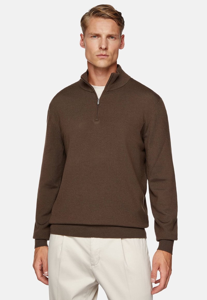 Brown zip-up sweater with ribbed texture and a high collar. Features long sleeves and a relaxed fit, paired with light-colored trousers.