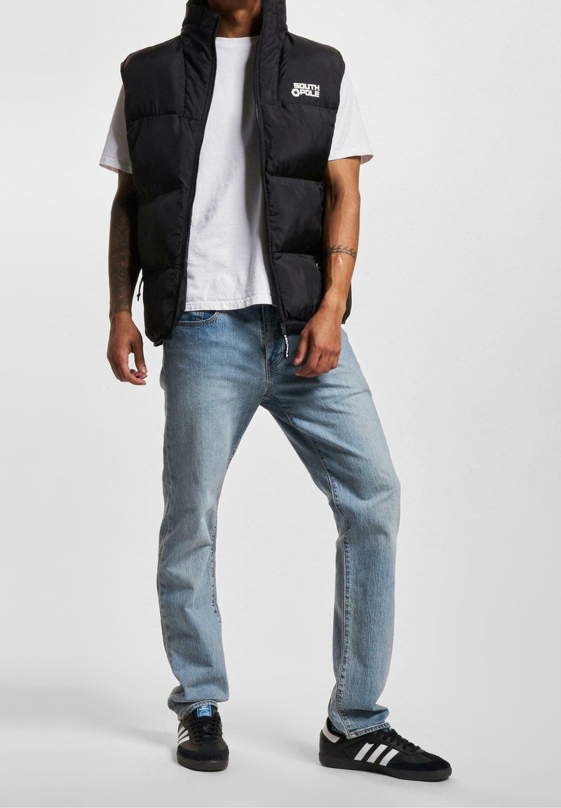 Personne portant une veste matelassée noire par-dessus un t-shirt blanc, un jean bleu clair et des baskets noires avec des rayures blanches, debout devant un fond uni.