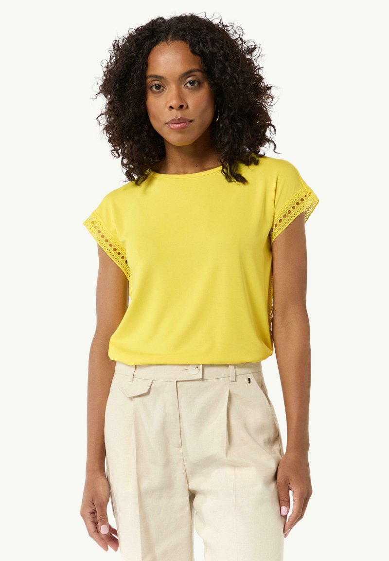 Yellow short-sleeve top with a smooth texture and lace detailing at the sleeves, paired with light beige trousers.