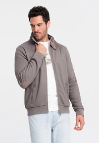 Giacca grigia con zip e finitura testurizzata, colletto marrone a contrasto e toppe sui gomiti, presenta due tasche laterali e un design aderente.