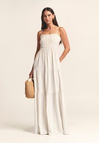 Bela maksi obleka z nabranim zgornjim delom, tankimi naramnicami in večplastno krilom z nežnimi navpičnimi črtami; model z rokah drži pleteno slamnato torbo.