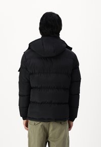 Schwarze Daunenjacke mit Kapuze, ausgestattet mit horizontalen Steppnähten und elastischen Bündchen, getragen über grünen Hosen. Die Rückansicht zeigt die Textur.