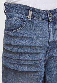 Gros plan sur un jean en denim bleu avec poche avant, passant de ceinture, bouton en métal et détails de plis horizontaux au niveau de la cuisse.