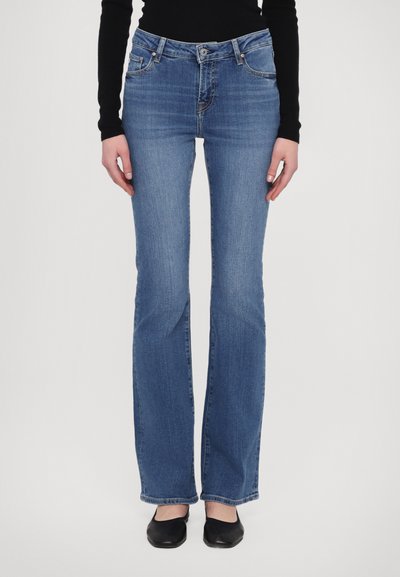 Blå flagrende jeans i denim med mellemhøj talje, der har frontlommer, en enkelt knaplukning og subtile falmede områder. Sorte flade sko nedenfor.