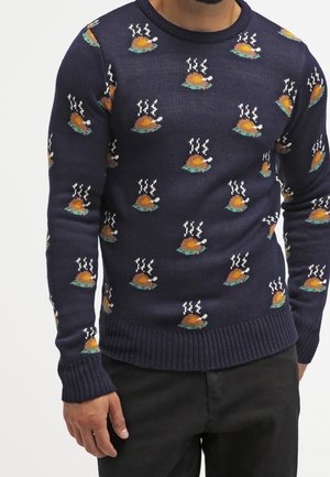 Granatowy sweter z powtarzającym się wzorem gotującego się pieczonego indyka, noszony przez osobę w czarnych spodniach, pokazany od szyi do ud.