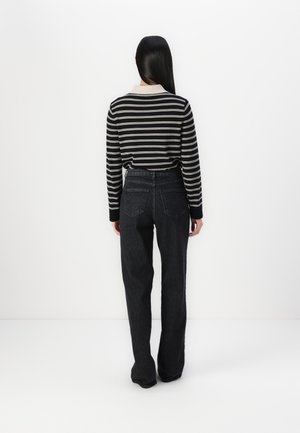 Frau mit langen schwarzen Haaren, die ein schwarz-weiß gestreiftes Langarmshirt und hoch taillierte schwarze Jeans trägt, steht einer weißen Wand gegenüber.