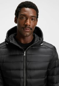 Giacca a vento nera con chiusura zip frontale, motivo a cuciture orizzontali e cappuccio. Presenta accessori in metallo argento e una texture liscia e lucida.