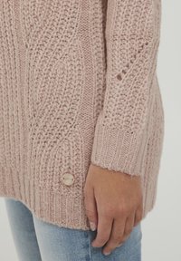 Ljusrosa stickad tröja med strukturerade kabelmönster, ribbade ärmslut och en dekorativ knappdetalj vid nederkanten.