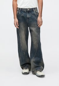 Hlače iz denim tkanine z širokimi hlačnicami v temno, posvetljenem modrem odtenku z vidnim nagubanjem. Imajo klasičen dizajn s petimi žepi in zapenjanje s pritrdilnim gumbom v pasu.