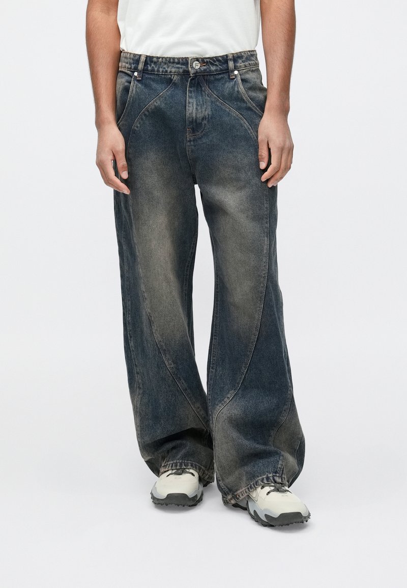 Hlače iz denim tkanine z širokimi hlačnicami v temno, posvetljenem modrem odtenku z vidnim nagubanjem. Imajo klasičen dizajn s petimi žepi in zapenjanje s pritrdilnim gumbom v pasu.