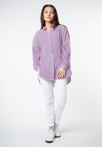 Helllila Button-up Hemd aus strukturiertem Stoff; kombiniert mit weißen Hosen und weißen Turnschuhen. Das Hemd hat einen lässigen Schnitt und einen längeren Rücken.