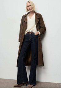 Abrigo largo de ante marrón sobre un suéter de punto blanco y suave, combinado con jeans oscuros acampanados y tacones marrones, con un cinturón ajustado.