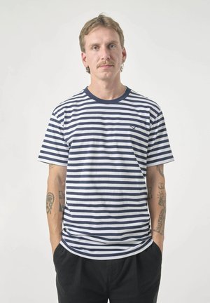 Man with light brown hair wearing a navy and white striped t-shirt and black pants, standing with hands in pockets against white background.