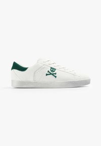 Zapatillas de ante blancas con detalles en ante verde, que presentan un bordado de calavera y huesos cruzados en el lateral, cordones rosas y una suela de goma texturizada.