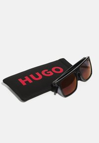 Occhiali da sole neri con lenti marroni e montatura quadrata, accompagnati da una custodia nera con il logo rosso "HUGO" in evidenza.
