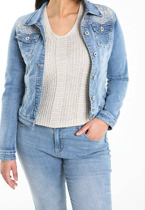 Femme portant une veste en denim bleu clair avec broderies sur les épaules, un pull en maille crème et un jean bleu clair, la main dans la poche.