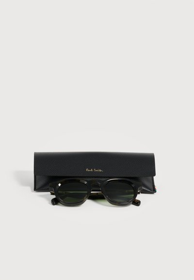Lunettes de soleil noires à monture épaisse avec des verres foncés reposant devant un étui noir texturé avec le logo Paul Smith doré sur fond blanc.