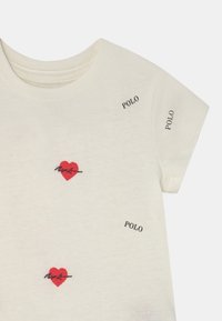 Krémové bavlněné tričko s krátkými rukávy, zdobené červenou výšivkou srdce a slovem "POLO" vytištěným černě kolem designu.