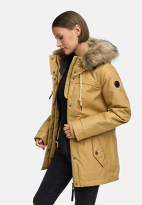 Gul parka-jakke med falsk pelshette, glidelås og trykknapper, sidelommer og snor ved halsen, brukt over en svart topp og jeans.