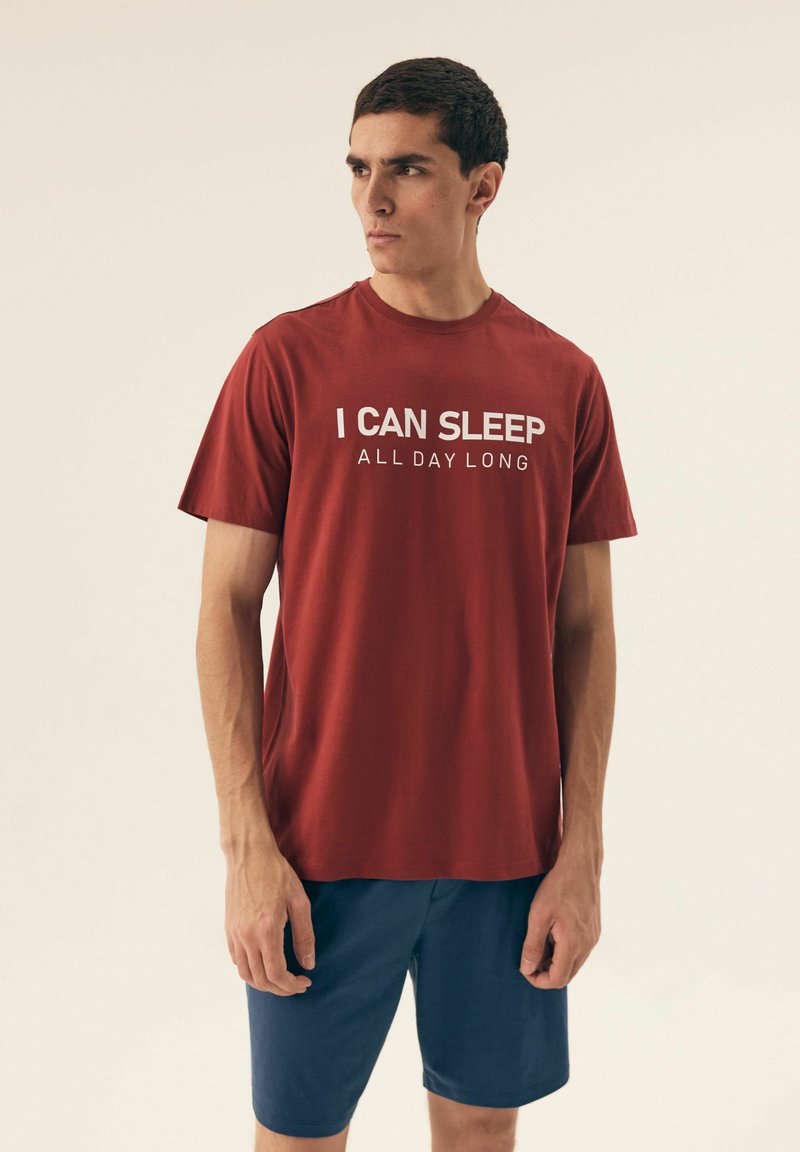Czerwona bawełniana koszulka z krótkim rękawem i okrągłym dekoltem, z białym napisem: "MOGĘ SPAĆ CAŁY DZIEŃ." Połączona z granatowymi szortami.