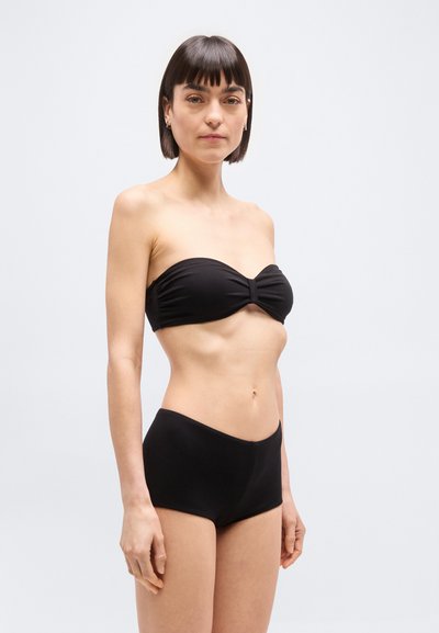 Γυναίκα με κοντά σκούρα μαλλιά, που φοράει μαύρο μπικίνι τοπ χωρίς λουριά και μαγιό με ψηλή μέση, στέκεται μπροστά σε απλό ανοιχτόφωτο φόντο.