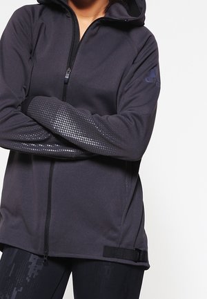 Schwarze Zip-Hoodie mit Kapuze, die texturierte Akzente an den Ärmeln und ein Logo auf der Schulter aufweist. Hergestellt aus glattem, dehnbarem Material.