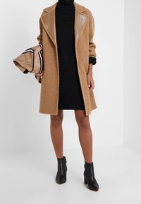 Abrigo marrón texturizado con solapas brillantes marrones, combinado con un vestido negro de cuello alto. Los accesorios incluyen un bolso beige a rayas y botines negros.