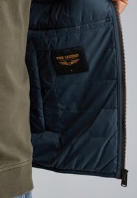 Marinblå isolerad jacka med en slät yta, prydd med en PME Legend-etikett i svart och orange. Sidog zip med en robust dragflik.