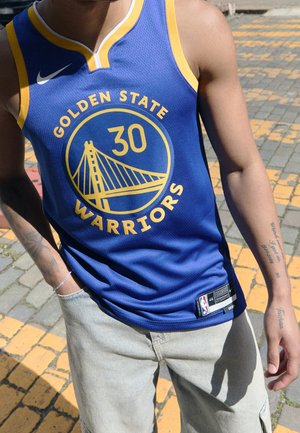 Person trägt ein blaues Golden State Warriors Basketballtrikot mit der Nummer 30 und hellen Hosen, steht auf einer gepflasterten Fläche.