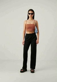 Schwarze weit geschnittene Hose kombiniert mit einem gemusterten Cropped-Top in Orange und Rot, mit dünnen Trägern. Die Accessoires umfassen eine kleine blaue Tasche und Sonnenbrille.
