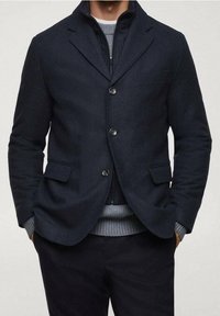 Man in een gelaagde outfit met een donkerblauwe blazer over een zwarte zip-up jas en een grijze gebreide trui, met zijn handen in zijn zakken.