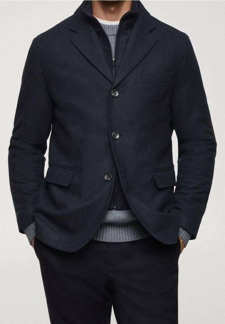 Man in een gelaagde outfit met een donkerblauwe blazer over een zwarte zip-up jas en een grijze gebreide trui, met zijn handen in zijn zakken.