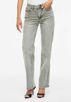 Kvinde iført lysegrå jeans med lige ben og høj talje samt sorte højhælede sandaler med åben tå, poserer med den ene hånd i lommen.