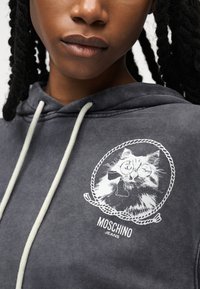 Κοντινό πλάνο προσώπου που φορά γκρι φούτερ με λευκά κορδόνια και εκτυπωμένη γάτα με γυαλιά και παπιγιόν μέσα σε πλαίσιο σχοινιού.