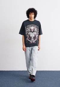 Svart grafisk t-skjorte med intrikat design som viser en menneskelig figur, kombinert med lyseblå smale jeans og svarte joggesko.